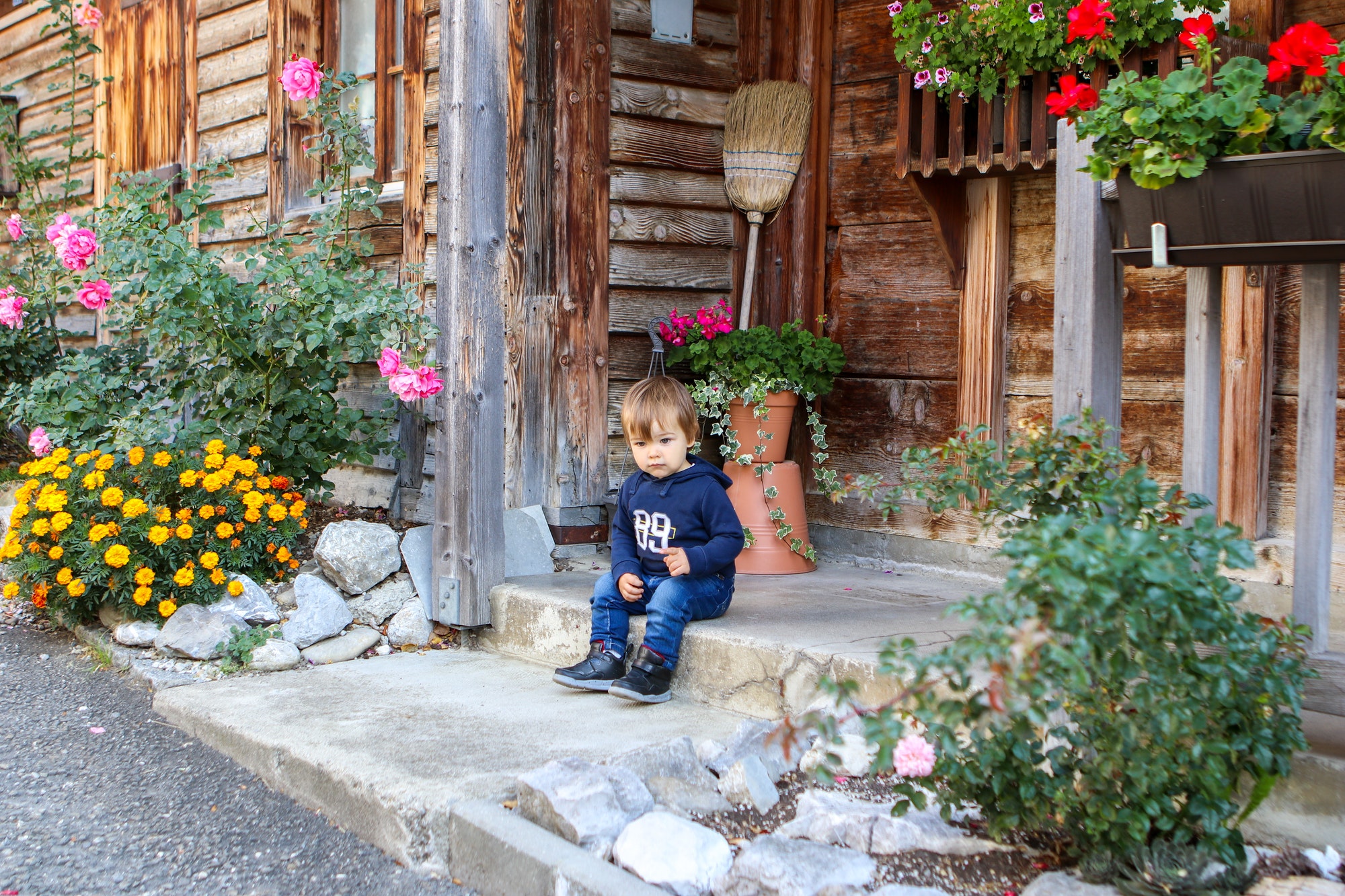 Cute little thoughtful boy sitting on the front porch of old vintage wooden house surrounded by